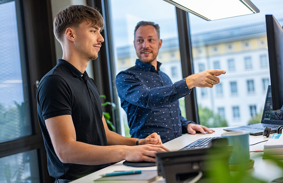 Dualer Student steht mit einem Kollegen am Schreibtisch und lässt sich etwas erklären