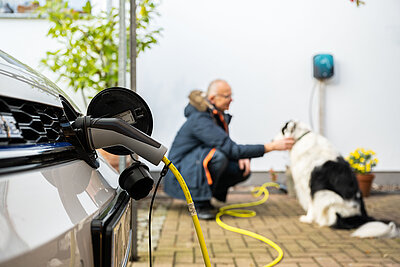 Ein Elektroauto lädt, der Stecker steckt in der Ladebuchse. Im Hintergrund streichelt ein Mann seinen Hund, ebenso ist die SMW Wallbox an der Hauswand zu erkennen.