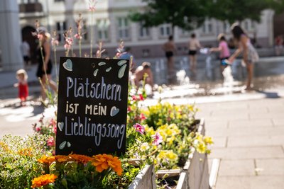 Beetschild "Plätschern ist mein Lieblingssong" in Nachbars Garten mit Wasserspiel im Hintergrund