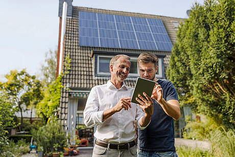 Zwei Männer stehen vor einem Haus mit PV Anlage und schauen auf ein Tablet