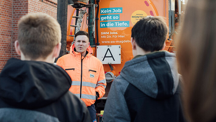 Mitarbeiter von SWM erklärt Schülern den Arbeitsalltag bei SWM - im Hintergrund ist das Kanalspülfahrzeug zu sehen